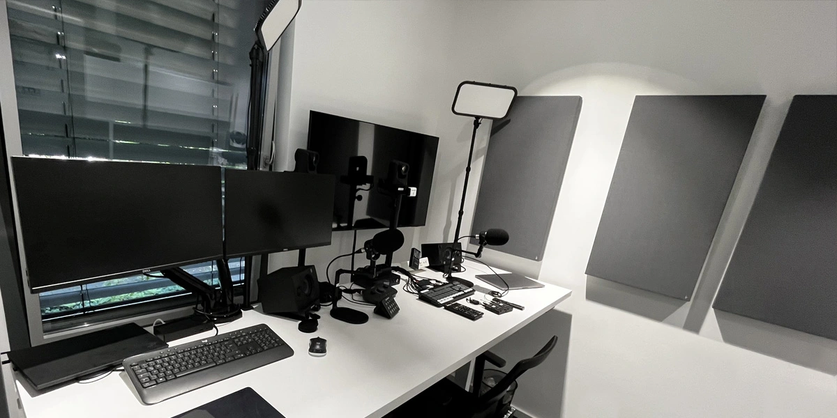corporatestudio_001 Ein moderner Arbeitsbereich mit drei Monitoren, einer Tastatur, einer Maus, einem Mikrofon, professioneller Beleuchtung, Kameras und anderen audiovisuellen Geräten auf einem weißen Schreibtisch. An der angrenzenden Wand sind Akustikplatten angebracht.