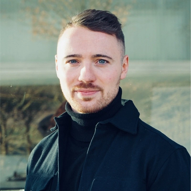 Vincent Höppner Ein Mann mit kurzen braunen Haaren und gestutztem Bart trägt einen schwarzen Rollkragenpullover und eine Jacke. Er steht im Freien in weichem natürlichem Licht und schaut mit einem leichten Lächeln direkt in die Kamera.