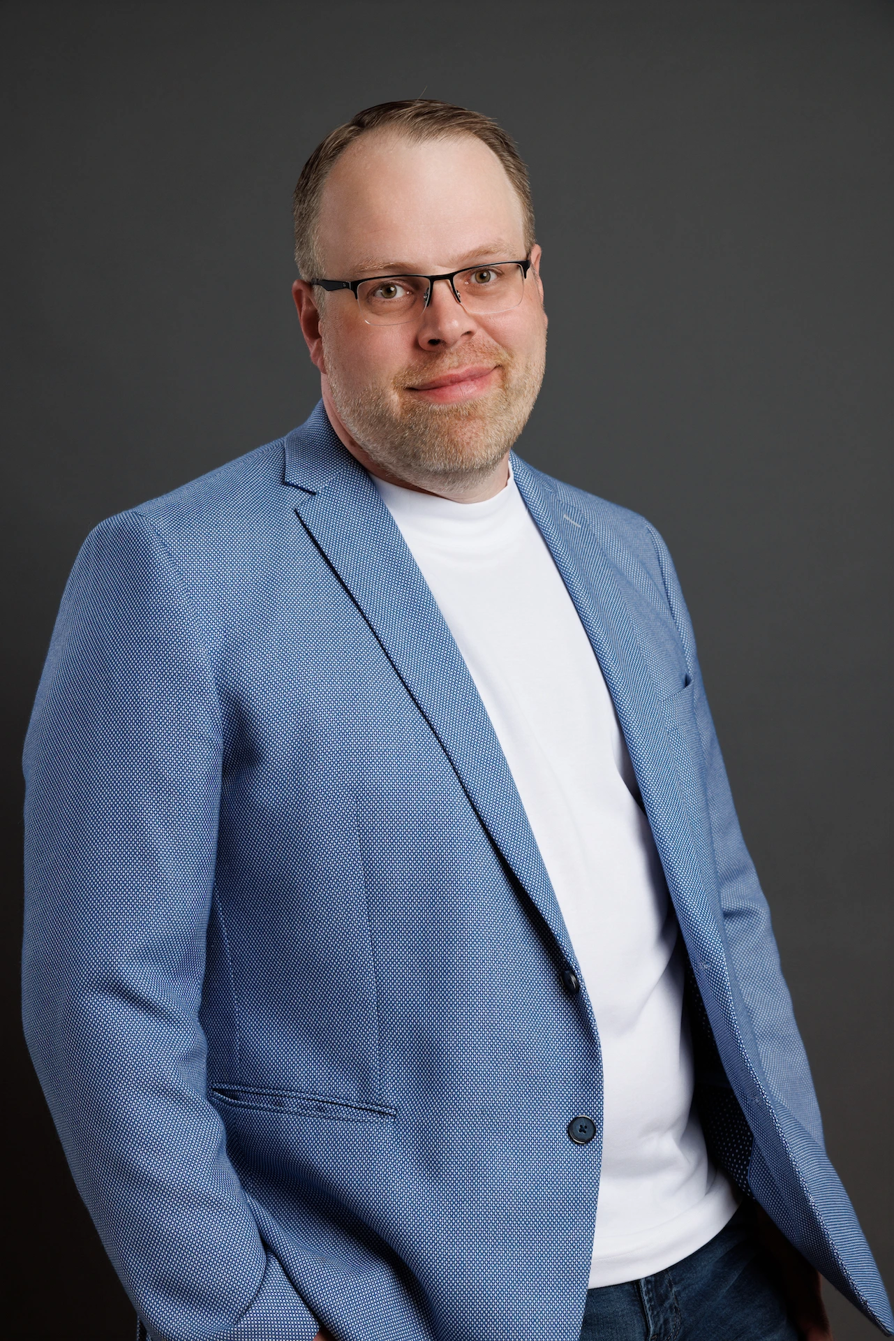 Florian_Business_44 Ein Mann mit kurzen hellen Haaren und Brille, der einen hellblauen Blazer, ein weißes T-Shirt und Jeans trägt, steht vor einem schlichten dunklen Hintergrund und lächelt leicht in die Kamera.