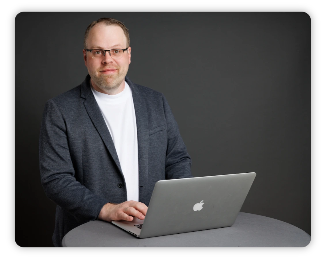 Florian_Homepage_schatten Ein Mann, der eine Brille, ein weißes Hemd und einen dunklen Blazer trägt, steht an einem runden Tisch und tippt auf einem silbernen Apple-Laptop. Der Hintergrund ist schlicht und dunkelgrau.