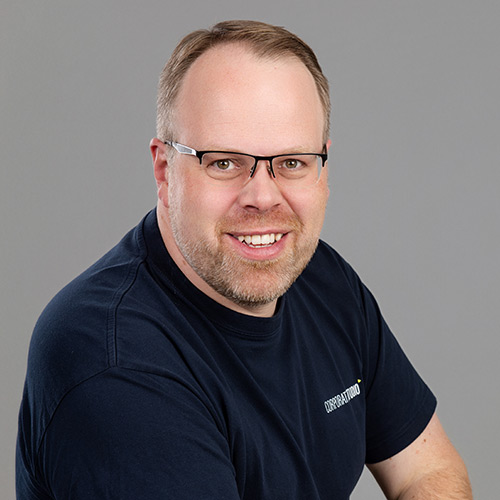 Ein lächelnder Mann mit kurzen hellbraunen Haaren, Brille und einem kurzen Bart trägt ein marineblaues T-Shirt und posiert vor einem schlichten grauen Hintergrund.