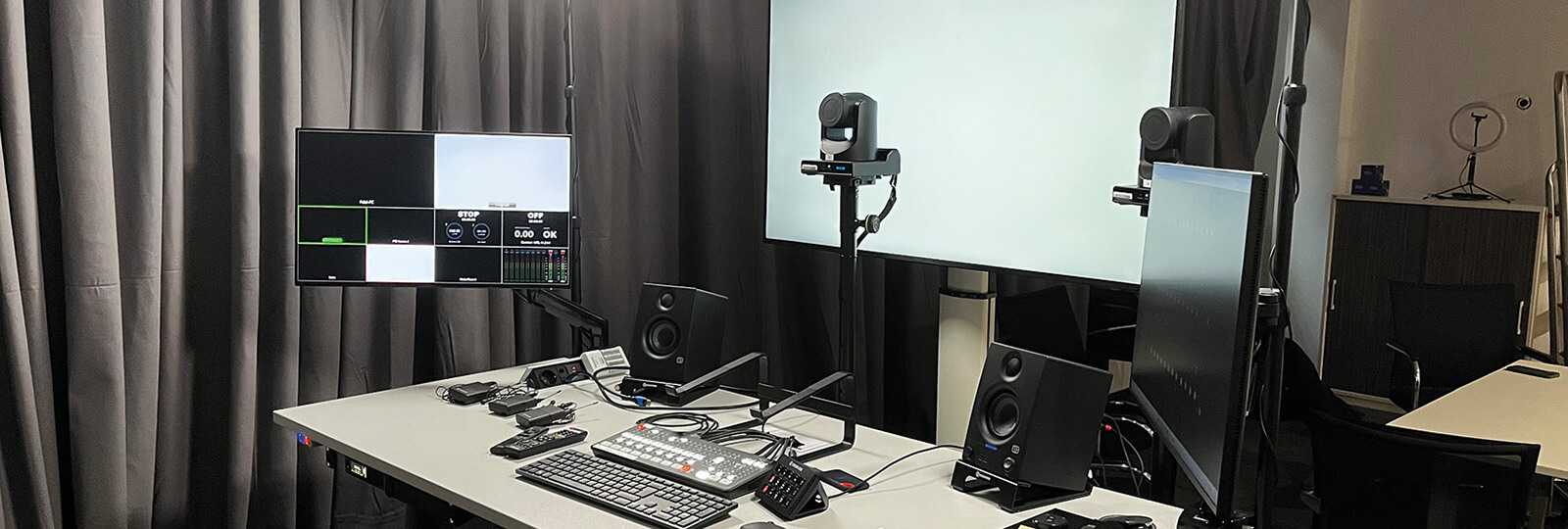 Ein modernes Videoproduktionsstudio mit Kameras, Monitoren, Mikrofonen, Lautsprechern und Steuerpulten, die auf Tischen vor schwarzen Vorhängen und einem großen weißen Hintergrund aufgebaut sind.
