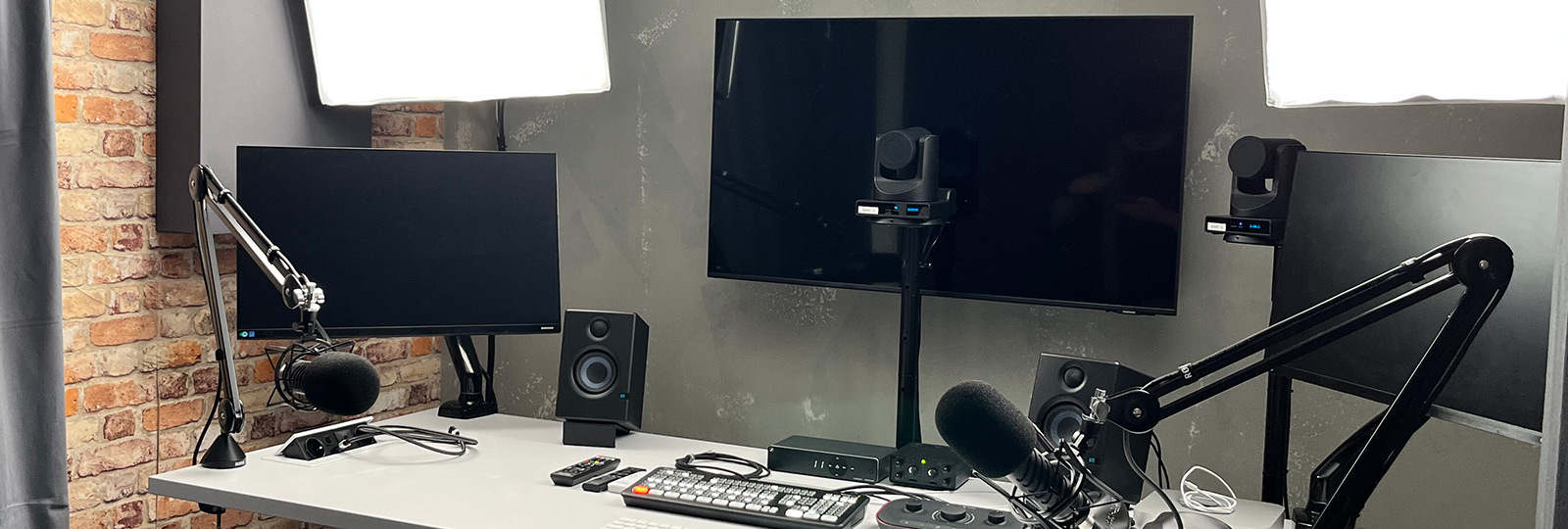 Ein modernes Podcast- oder Aufnahmestudio mit zwei Mikrofonen an verstellbaren Armen, Lautsprechern, zwei Monitoren, Kameras, Studiolicht und einem Steuerpult auf einem weißen Schreibtisch vor einer ziegelgrauen Wand.