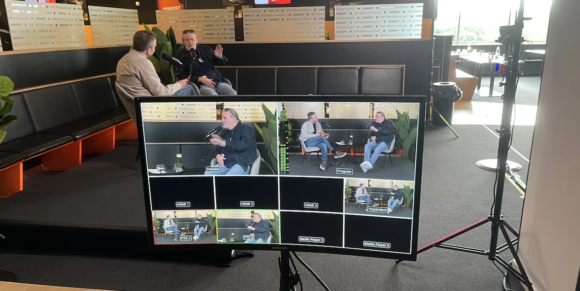 Auf einem Kameramonitor sind mehrere Live-Videobilder von zwei Personen zu sehen, die sich in einem modernen Innenraum unterhalten. Der Monitor befindet sich im Vordergrund, während das eigentliche Gespräch im Hintergrund stattfindet.
