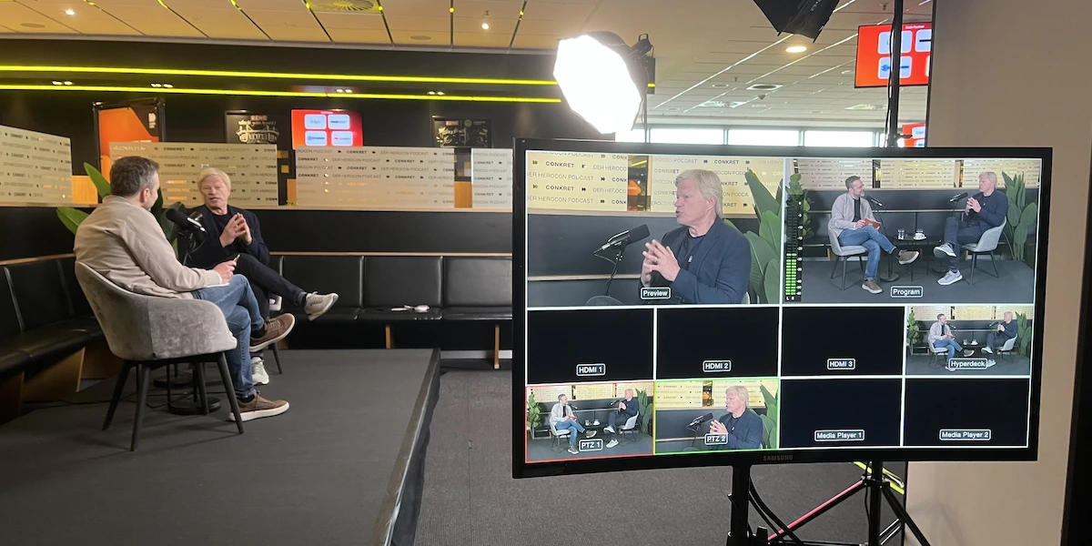 Zwei Personen sitzen auf einer Bühne in einem Studio mit professioneller Beleuchtung und unterhalten sich; auf einem Monitor in der Nähe sind verschiedene Kameraeinstellungen ihres Gesprächs zu sehen.