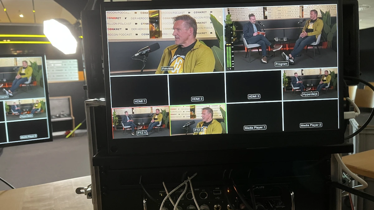 Auf einem Videoproduktionsmonitor sind mehrere Kameraeinstellungen von zwei Männern zu sehen, die in einem Studio einen Podcast aufnehmen. Ein Mann spricht in ein Mikrofon, während der andere zuhört. Unterhalb des Monitors sind Kabel und Geräte zu sehen.